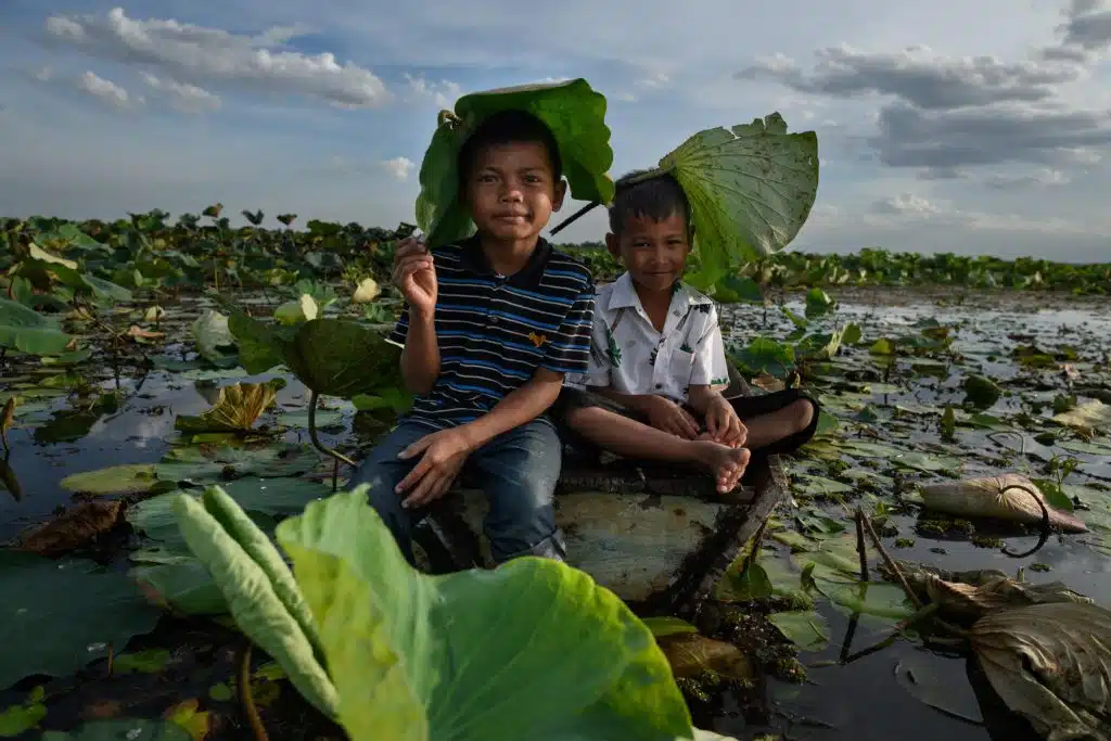 (À droite) Taweesap Panthong (Kao) et (à gauche) Siwara Trongsundee (Fai) sont assis sur une surface plane entourée de grandes feuilles de lotus dans un plan d’eau. C’était une journée d’été très chaude en Thaïlande, en mai, où la canicule rendait les activités en extérieur presque impossibles pour de nombreux enfants.